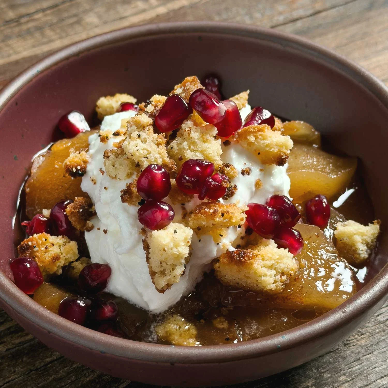 Apple crumble cobbler topped with whipped cream and pomegranate seeds in a brown bowl.