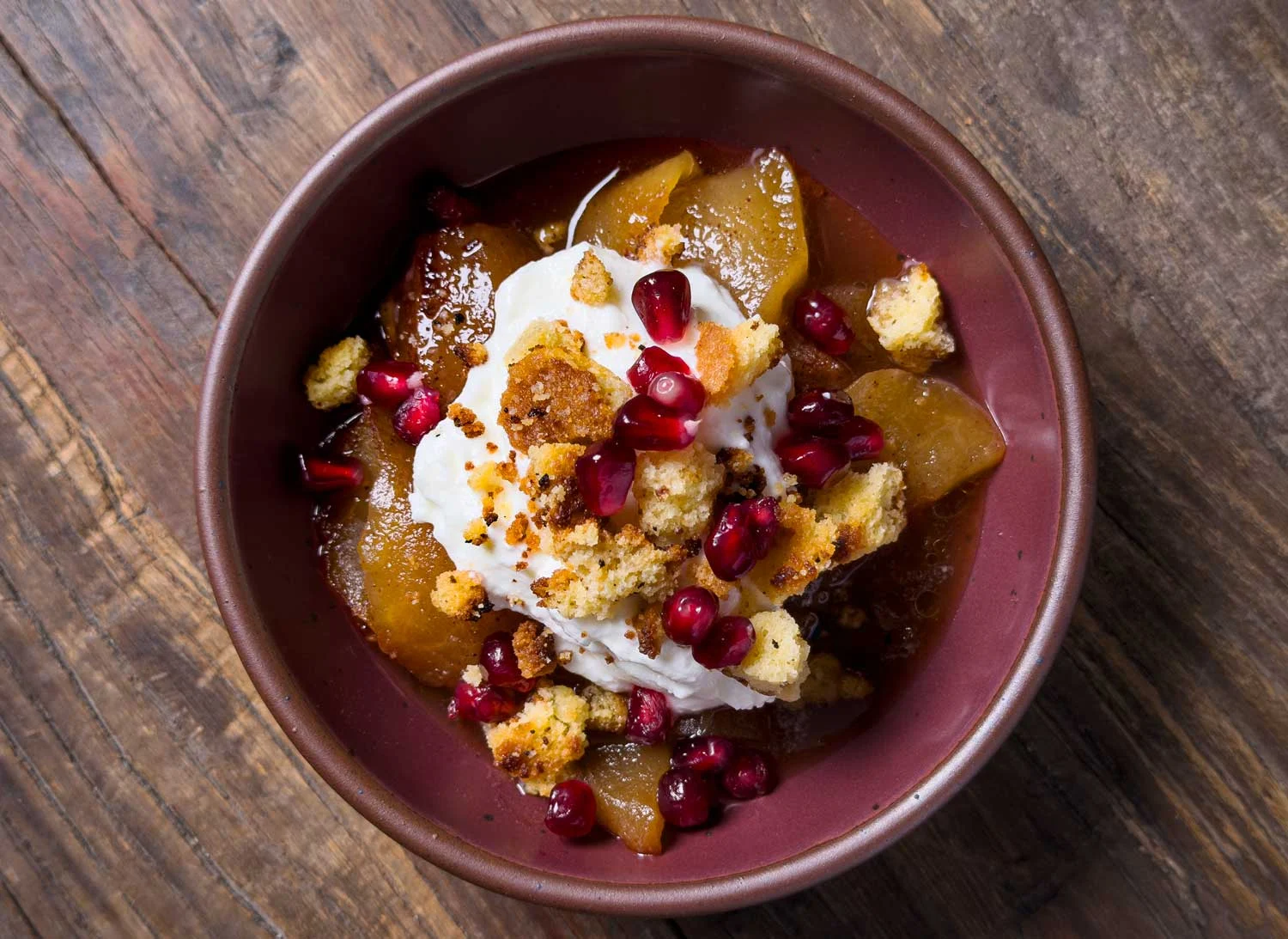Dessert bowl with poached pears in caramel sauce, topped with whipped cream, pomegranate seeds, and crumbly streusel pieces in a brown bowl on a wooden table