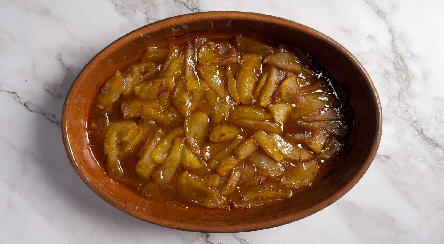 Oval clay dish filled with caramelized potato wedges in brown sauce on a white marble surface.