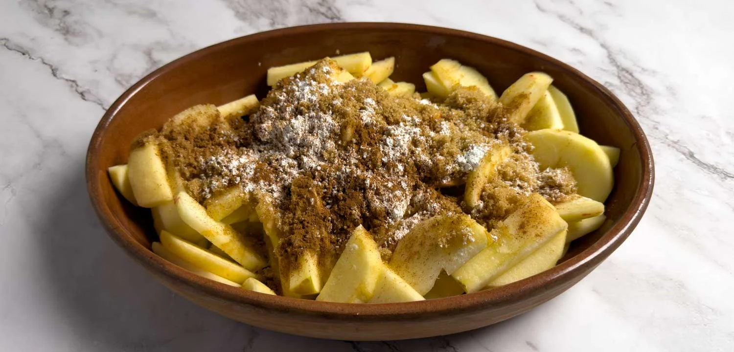 Brown ceramic bowl filled with sliced apples topped with cinnamon-spice crumbs and a light dusting of powdered sugar, on a marble surface.