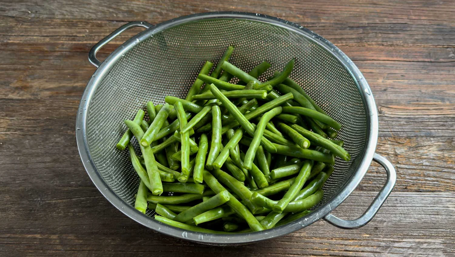 Fresh green beans in a metal colander on a wooden table, ready to cook