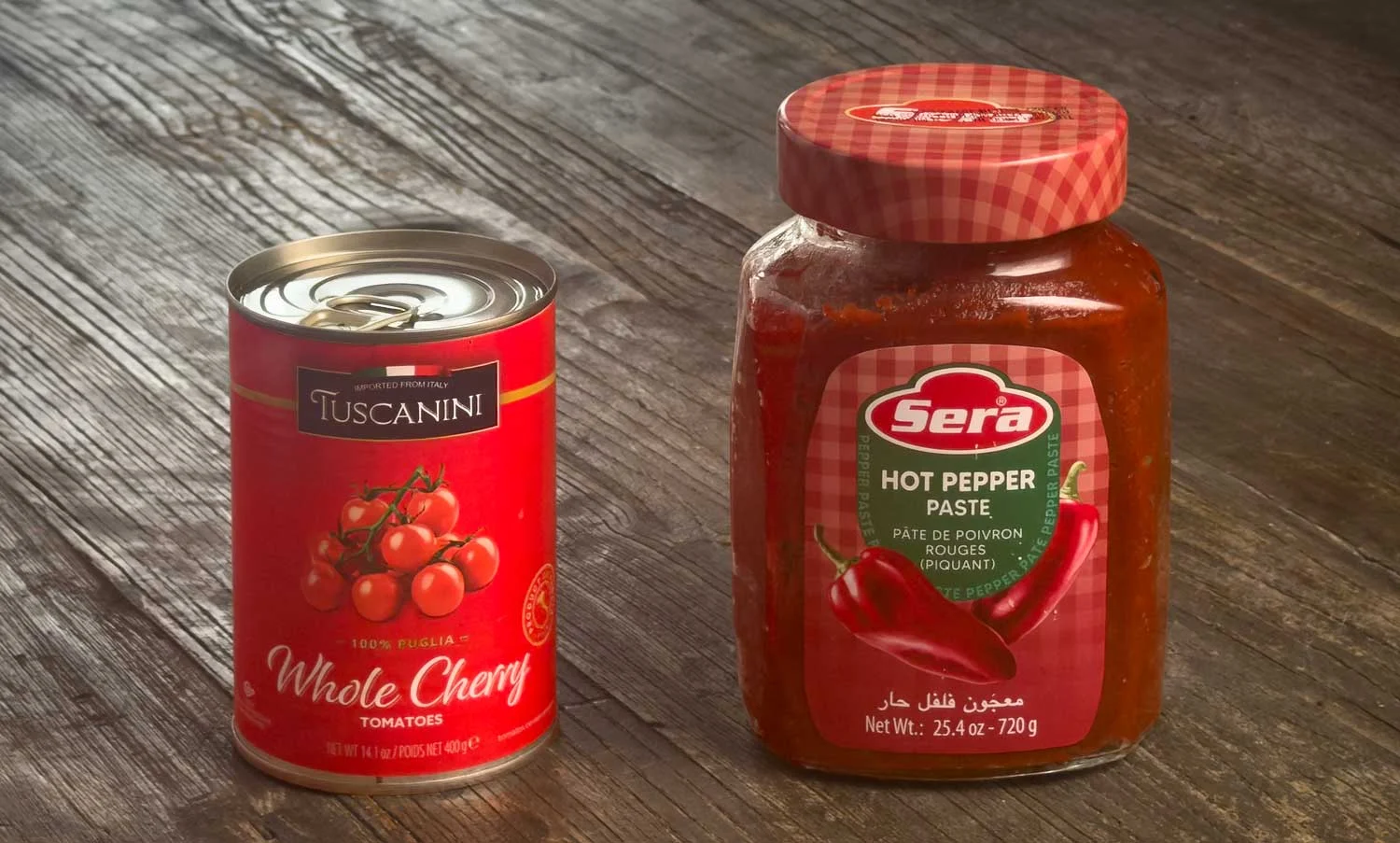 A red can of Tuscanini Whole Cherry Tomatoes next to a jar of Sera hot pepper paste on a wooden table.