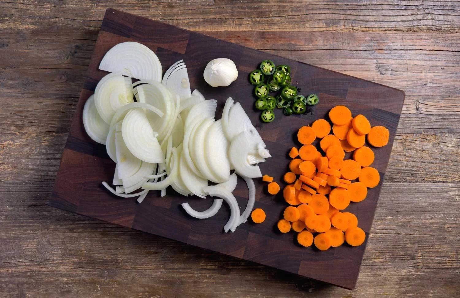 Sliced white onions in piles, orange carrot coins, and sliced jalapeños on a dark wooden cutting board with a garlic bulb nearby.